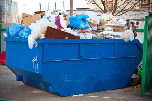 A large, blue industrial skip filled with mixed waste materials, including cardboard boxes, plastic sheeting, and paper, situated outdoors on a paved surface. The skip has a sturdy metal construction with a slightly textured finish, and the waste items are piled high, spilling slightly over the edges. Some of the waste, such as flattened cardboard and white plastic wrapping, is visible on the top, with other materials stacked underneath. In the background, there are faint outlines of trees and multi-storey buildings, indicating an urban or suburban environment. The setting suggests a commercial waste collection point, possibly in Stratford, where Commercial Waste Stratford provides rubbish removal services, with the skip positioned near a green fence or railing on the right side. Natural daylight highlights the textures of the waste and the weathered surface of the skip, emphasizing the practicality and purpose of waste management in a local town setting.