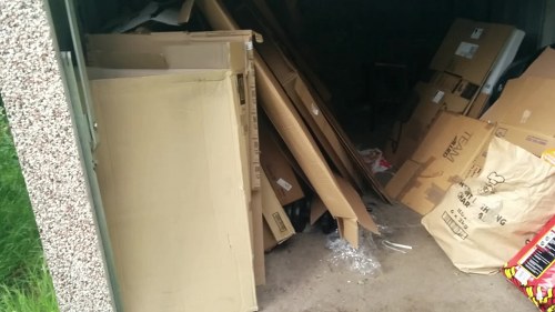 A collection of discarded office furniture and cardboard boxes arranged in a cluttered outdoor space, possibly a driveway or storage area. The items include a cream-colored wooden cabinet or shelf leaning against a wall, with textured surfaces. Surrounding this are various flattened cardboard boxes, some partially torn or crumpled, and a clear plastic bag with additional waste material. The background shows a dark, shadowed area with more cardboard and packaging materials stacked haphazardly, indicating reliance on rubbish removal services for clearing domestic or commercial waste. The scene is illuminated by natural light, highlighting the different textures of paper, wood, and plastic, and situating the image in a typical waste accumulation environment, relevant for local rubbish removal companies in Stratford.