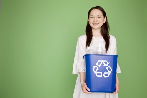A young woman with long dark hair, smiling and dressed in a white dress with lace sleeves, is standing in front of a plain green background. She is holding a blue recycling bin with a white recycling symbol on the front, positioned at waist level with both hands. The bin appears to be made of plastic, with a smooth surface and a slightly rounded top edge. The image is bright and well-lit, emphasizing the clarity of the woman's cheerful expression and the details of the recycling bin. This scene suggests a focus on waste separation and environmentally conscious practices, relevant to rubbish removal services offered by Commercial Waste Stratford in Stratford and surrounding areas, including the postcode district ST.