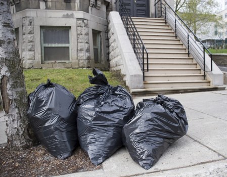 Three large black rubbish bags, made of plastic with a shiny finish, are placed on the concrete pavement next to a tree trunk on the left side of the image. The bags are full and tied at the top, indicating they contain collected waste. Behind the bags, there is a small patch of grass and a stone building with a rectangular window featuring a metal security grill. To the right, a set of external stairs with metal railings leads up to a building entrance. The area appears to be a residential or commercial frontage, with a clean, urban environment typical of Stratford. The scene is well-lit, suggesting daytime, and the overall setting emphasizes waste collection or removal activities, which would be relevant to services provided by Commercial Waste Stratford’s rubbish clearance offerings.